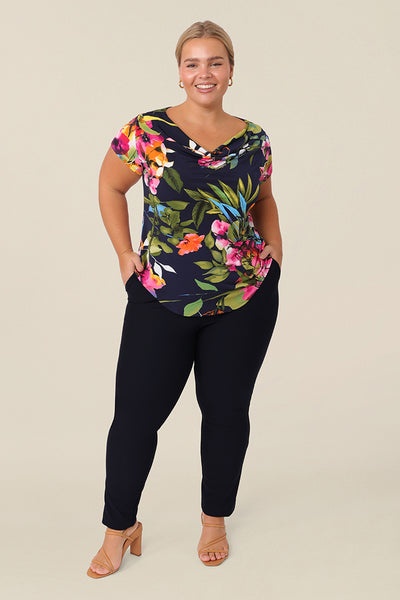 A size 14 curvy woman wears a cowl neck short sleeve jersey top in a floral print. She wears the top with work trousers in navy. Both are designed and made in Australia for petite to plus size women.