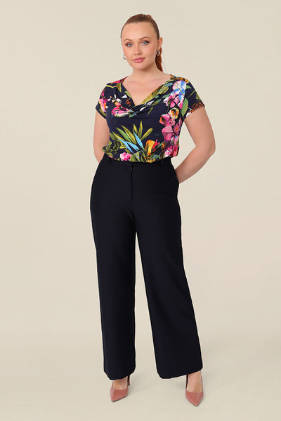 A size 12 curvy woman wears a cowl-neck short sleeve jersey top in a floral print. She wears the top with wide leg pants in navy. Both are designed and made in Australia for petite to plus size women.