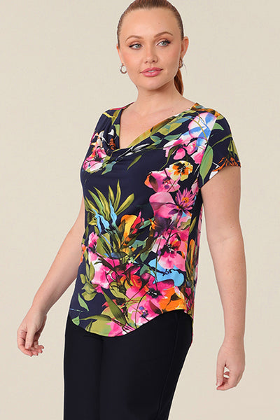 A size 12 curvy woman wears a cowl-neck short sleeve jersey top in a floral print. She wears the top with wide leg pants in navy. Both are designed and made in Australia for petite to plus size women.