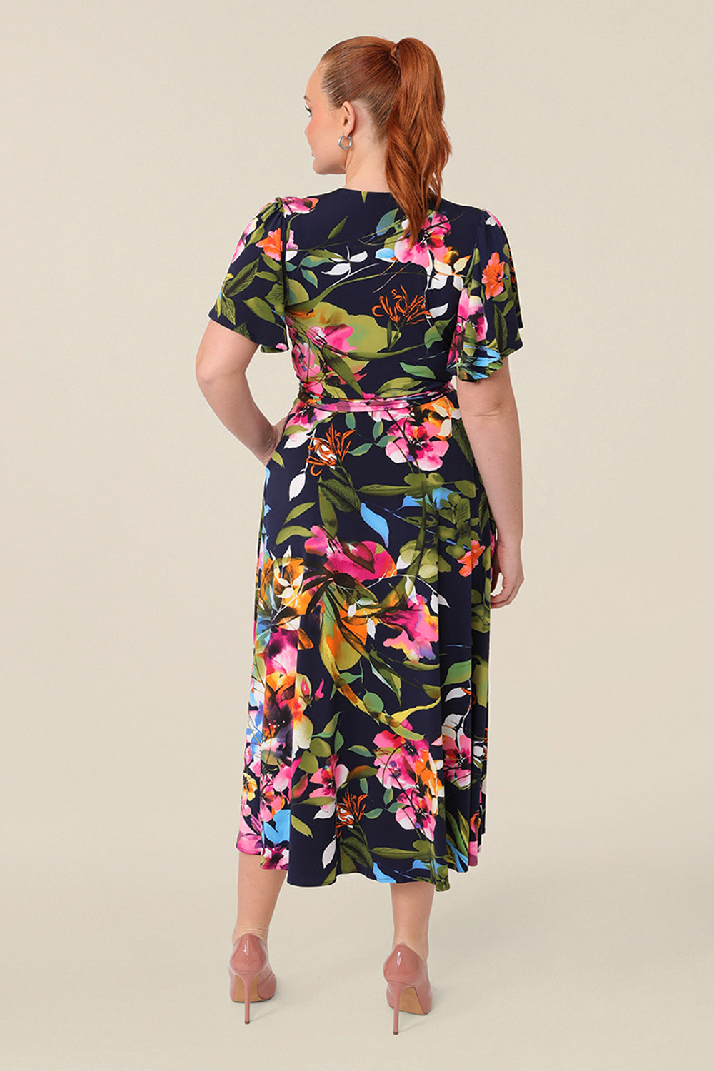 Back view of a A size 12 curvy woman wears a functioning wrap dress with peekaboo flutter sleeves. Featuring a midi-length skirt with wrap hem, this floral printed jersey dress is has a v-neckline and wears well as an everyday dress. 