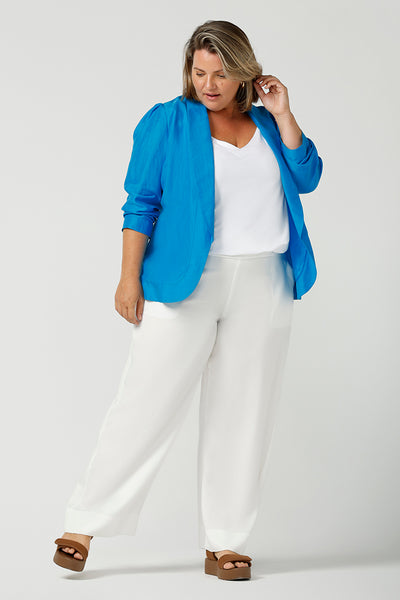 A curvy size, size 16 woman wearing a longline blazer in opal blue tencel fabric with a white bamboo cami and white woven pants. This lightweight jacket is comfortable for your everyday workwear, casual and travel capsule wardrobe. Shop this Australian-made blazer online in sizes 8 to 24, petite to plus sizes.