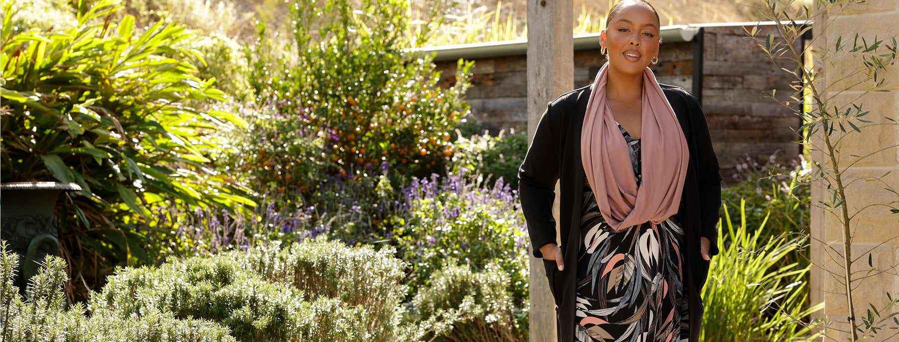a plus size woman shows how to do winter layering with a floral print jersey dress worn under a black coat and a pink bamboo jersey scarf. Shop the look at Australian and New Zealand women's clothing brand, Leina & Fleur.