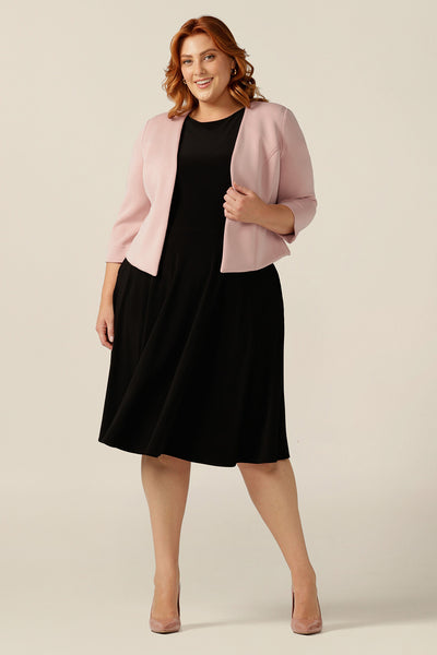 A woman wears a pink modal jacket with tailored panels, collarless neckline and 3/4 sleeves. Worn with a black work wear dress, this image shows how to add colour to corporate black dresses by shopping coloured work wear jackets
