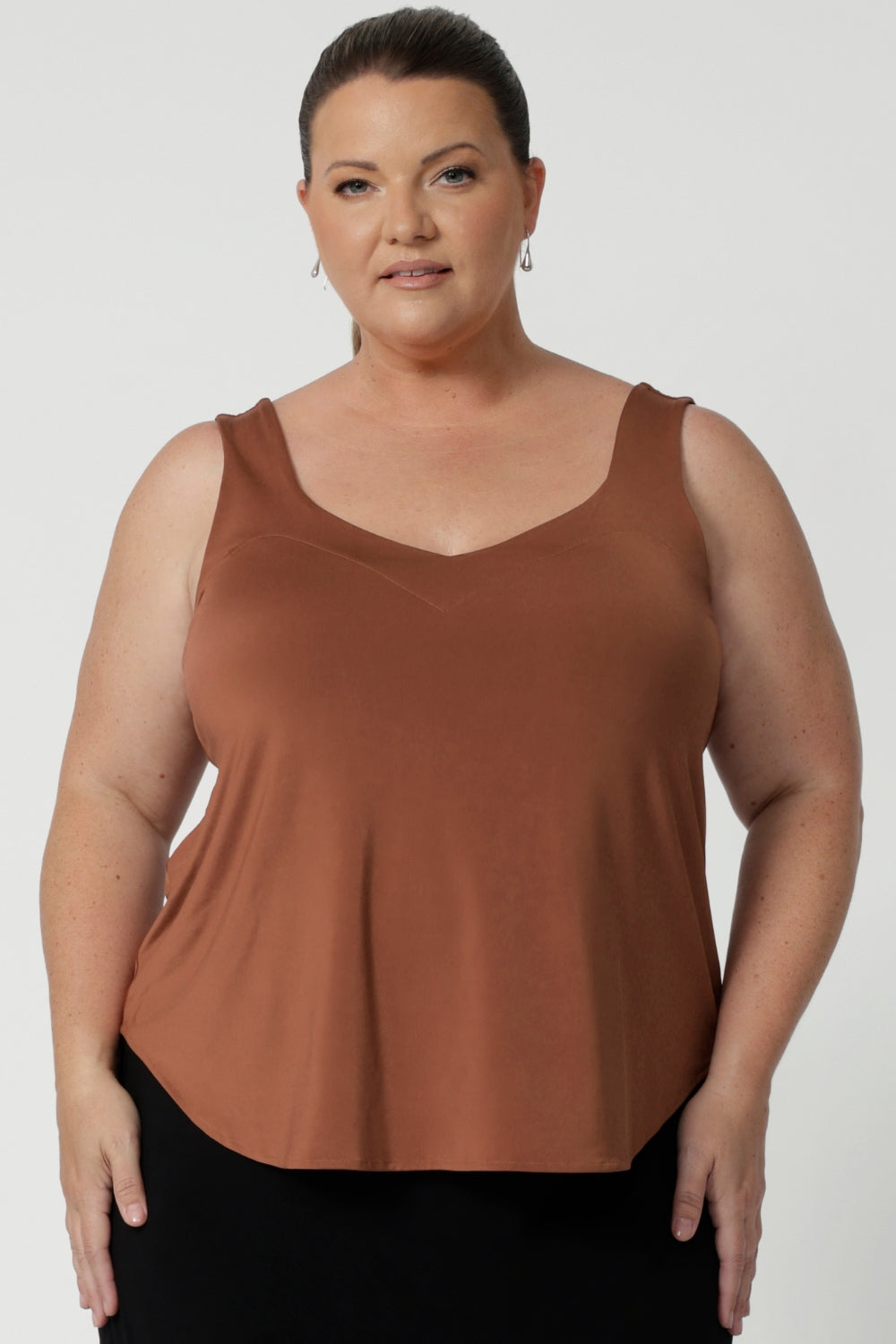 Plus sized woman wears clay coloured cami that features shirttail hemline, V cut at the front and back and wide shoulder straps. Crafted with care by Leina & Fleur, in slinky jersey fabric.