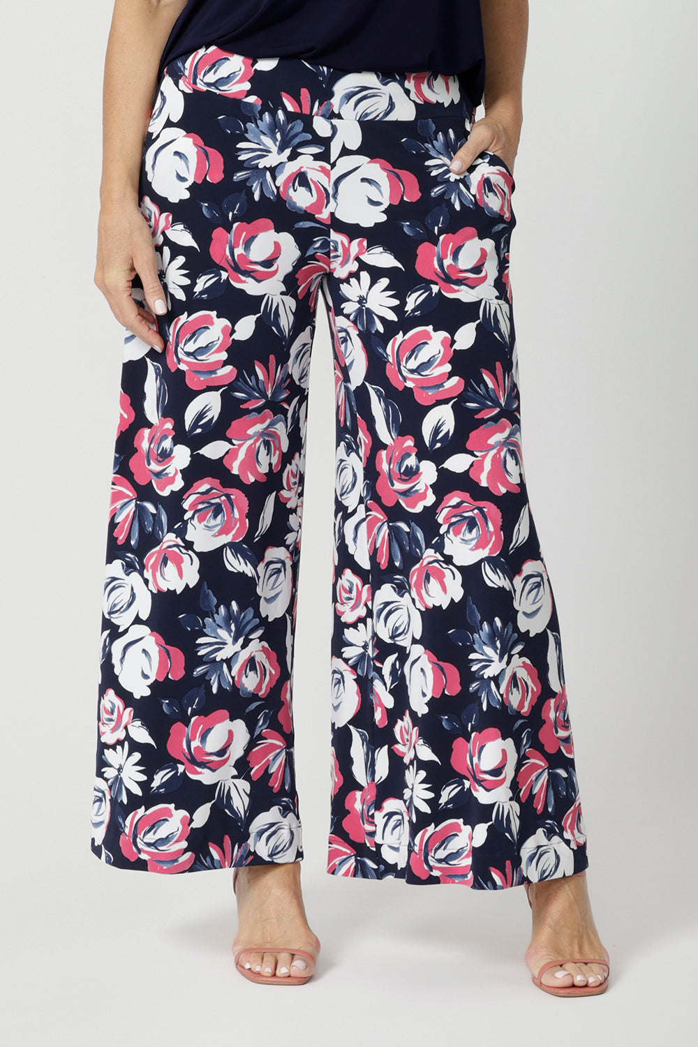 Close up front view of woman wearing wide legged pants. Floral printed pants are made of easy care dry touch jersey fabric. Colourful pants have a combination of pink and white roses on a navy pant. Pants are comfortable and perfect for travel. Fabric made in Australia by New Zealand and Australian women. Leina & Fleur is fashion brand that proudly stocks sizes from 8 to 24. Shop this fun and easy pant style in plus and petite sizes. 