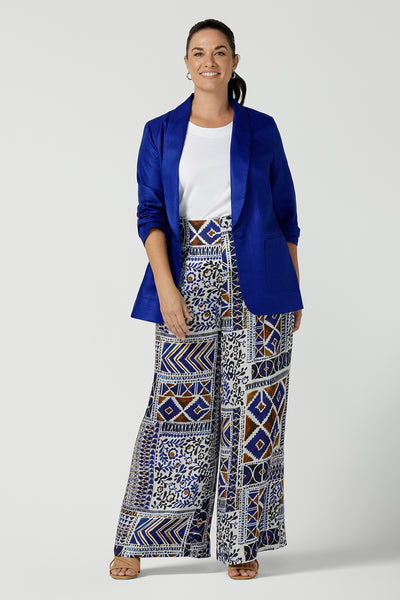 A size 12 woman wearing a Ellery pant in soft Italian Viscose fabric. Digitally printed in a Cadaques tile print. A pleat front style with self covered buttons and a wide leg opening and functioning pockets. Women wears a high waist satin wide leg pants in a Cadaques print. Styled back with the Houston Blazer in Cobalt Linen. Mad in Australia for women Size 8 - 24.