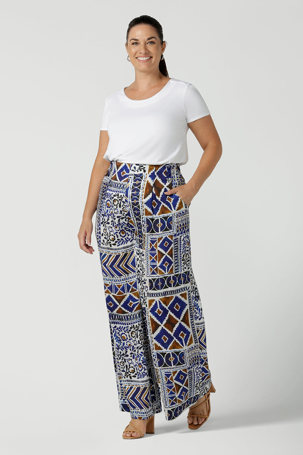 Size 12 woman wears a Ellery pant in soft Italian Viscose fabric. Digitally printed in a Cadaques tile print. A pleat front style with self covered buttons and a wide leg opening and functioning pockets. Women wears a high waist satin wide leg pants in a Cadaques print. Mad in Australia for women Size 8 - 24.
