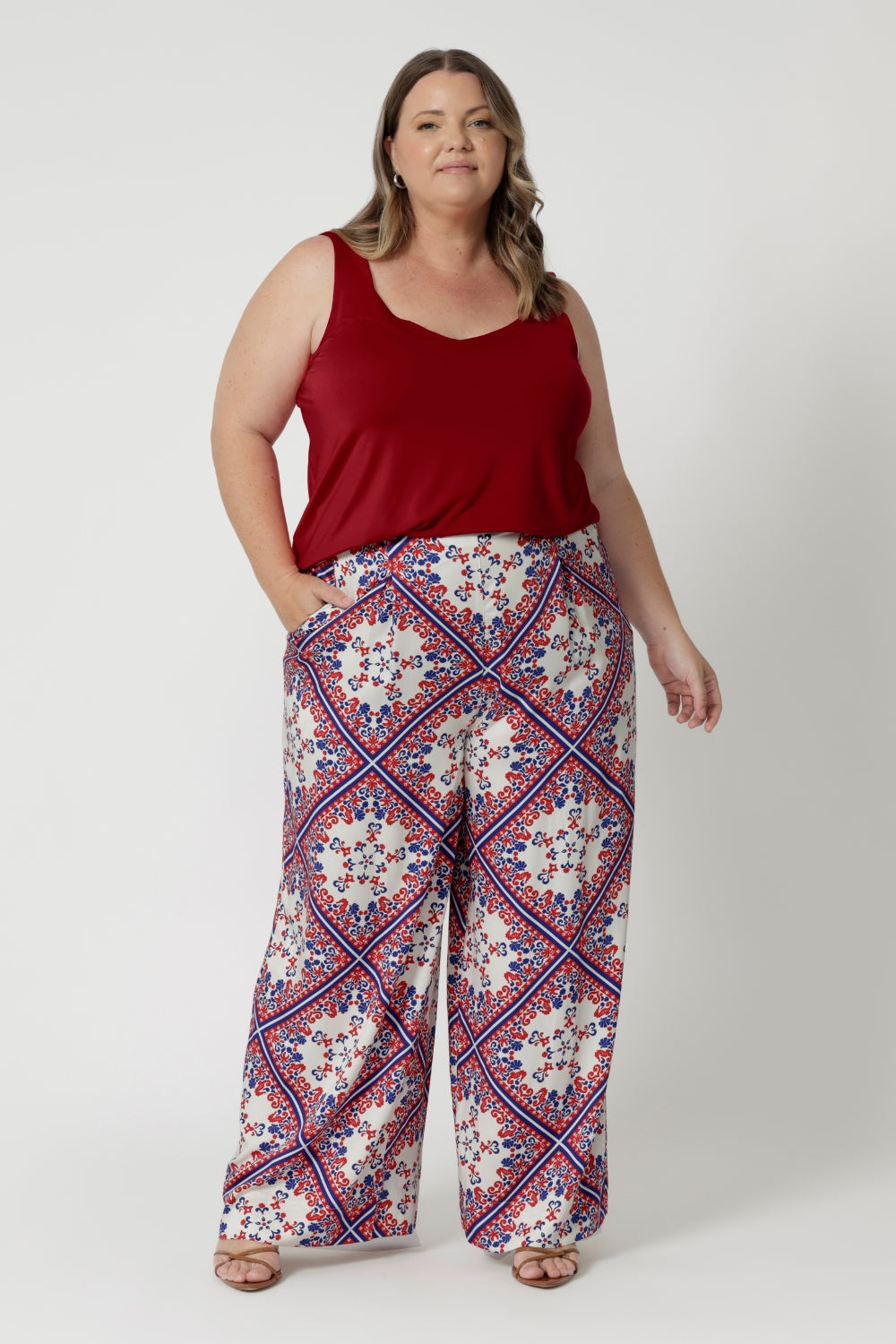 Plus sized woman wears Ellery pants in Seahorse print which displays a geometric print featuring a design mix of red & blue colours on a white base. Model pairs high waisted pants with red cami to tie the look together, matching the look with brown kitten heels. Pants available in size 8-24.