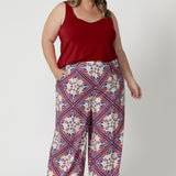 Plus sized woman wears Ellery pants in Seahorse print which displays a geometric print featuring a design mix of red & blue colours on a white base. Model pairs high waisted pants with red cami to tie the look together, matching the look with brown kitten heels. Pants available in size 8-24.