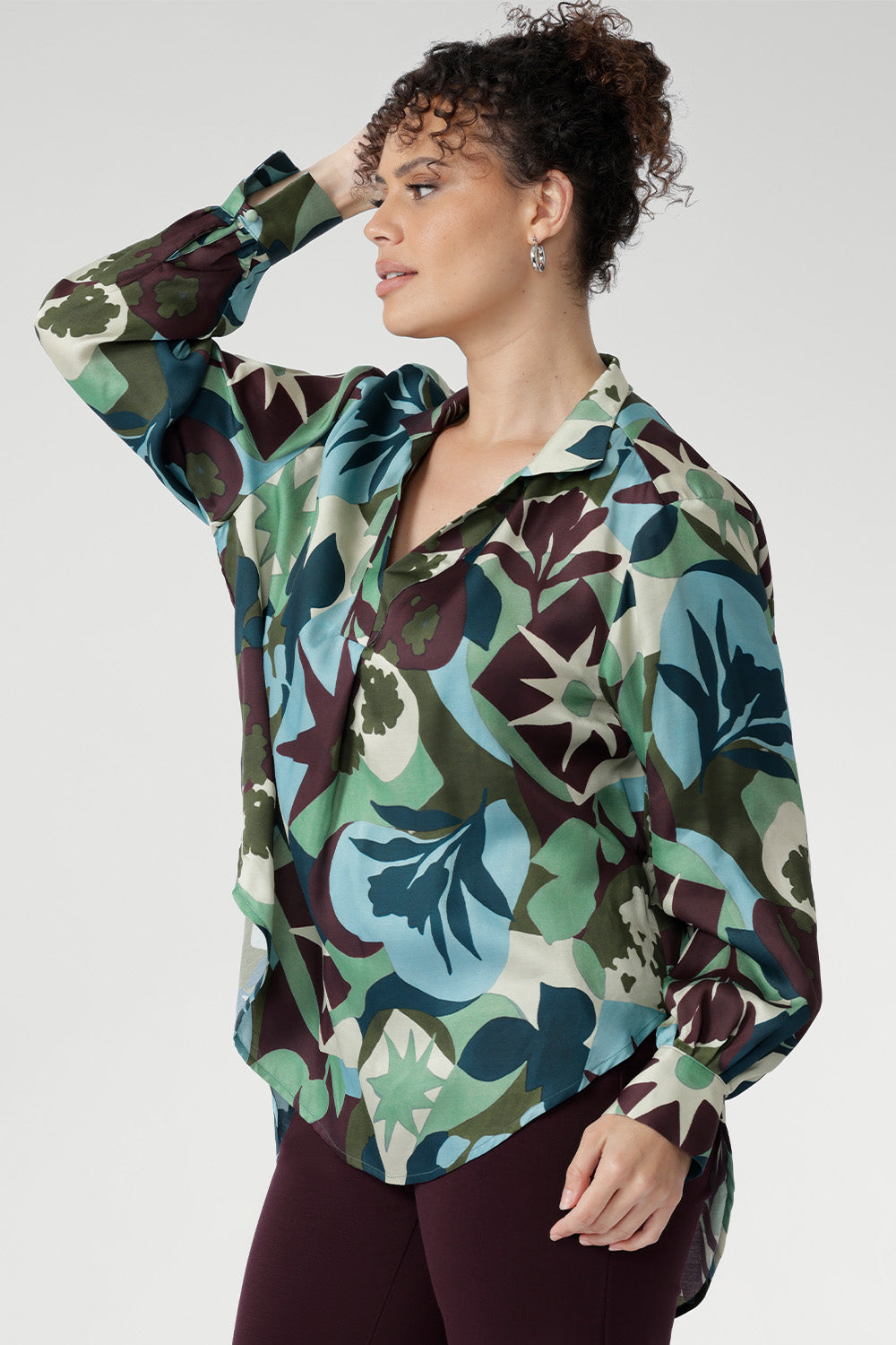 The bold abstract print blends rich tones of green, blue, and plum, making it a standout for both professional and social settings. A sophisticated collared V-neckline with tailored centre tuck adds structure, while the full-length cuffed sleeves with covered buttons bring a polished finish.