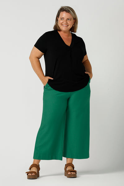 A good top for your capsule wardrobe, this V-neck black bamboo jersey top with short sleeves makes for a good office wear top for women, as well as a comfortable casual top. Worn with emerald green, wide leg tailored pants and shown on a size 18, curvy woman, this is a good top for plus size women. Made by Australian ladies clothing brand, Leina & Fleur, shop black tops for women in sizes 8 to 24 in their online fashion boutique.