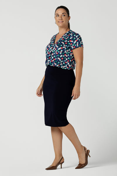 Front view of a happy corporate woman wearing the Emily Top in Blue bubbles spot print. Short sleeves with a pleat front. V-neckline and back yoke. Comfortable jersey fabric. Made in Australia for women size 8 - 24. Styled back with the black Andi skirt a comfortable tube work skirt.