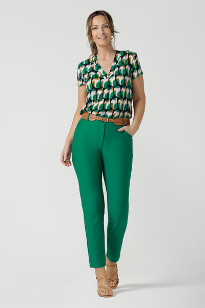 a size 10 woman wearing a v-neck short sleeve jersey top in a geometric print. She wears the top with emerald green tapered leg pants and a tan belt. Designed and made in Australia for petite to plus size women.
