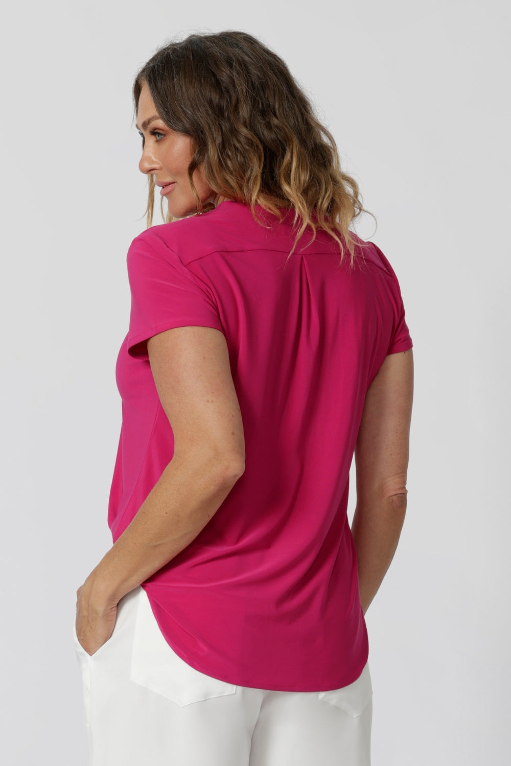 An back image of a woman in her forties wearing a hot pink work top with a v neck. Her top is made from a stretch jersey by fashion label Leina & Fleur. It is made in Australia in sizes 8 to 24.