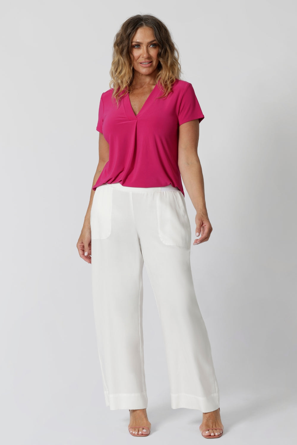 An image of a woman in her forties wearing a hot pink work top with a v neck and white pants made from sustainable lyocell fibres. Her top is made from a stretch jersey by fashion label Leina & Fleur. It is made in Australia in sizes 8 to 24.