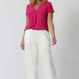 An image of a woman in her forties wearing a hot pink work top with a v neck and white pants made from sustainable lyocell fibres. Her top is made from a stretch jersey by fashion label Leina & Fleur. It is made in Australia in sizes 8 to 24.