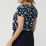 Back view of a good work top for women, a size 12, over 50s woman wears a short sleeve, V-neck top in aqua blue floral print. This stretch jersey top for work is worn tucked in to  Navy blue for pants for corporate outfit, Made in Australia by Australian and New Zealand women's clothing label, Leina & Fleur, shop over 50s fashion online in sizes 8 to 24.