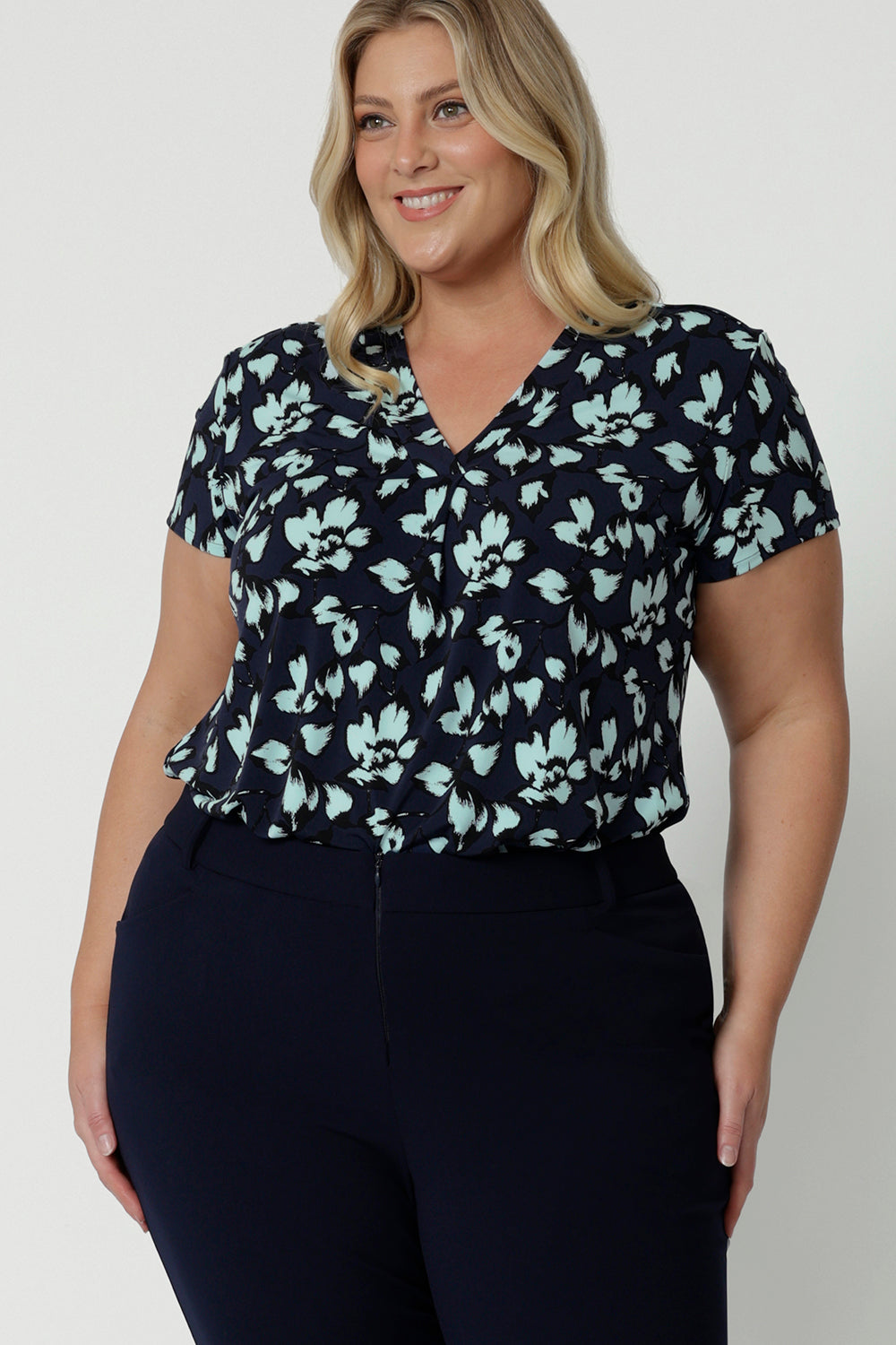 Showing a good top for plus size women, a size 18, curvy woman wears short sleeve, V-neck top in aqua blue floral print. This stretch jersey top for work is worn tucked in to Navy blue for pants for corporate outfit, Made in Australia by Australian and New Zealand women's clothes brand, Leina & Fleur, shop curve fashion online in sizes 8 to 24.