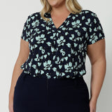 Showing a good top for plus size women, a size 18, curvy woman wears short sleeve, V-neck top in aqua blue floral print. This stretch jersey top for work is worn tucked in to Navy blue for pants for corporate outfit, Made in Australia by Australian and New Zealand women's clothes brand, Leina & Fleur, shop curve fashion online in sizes 8 to 24.