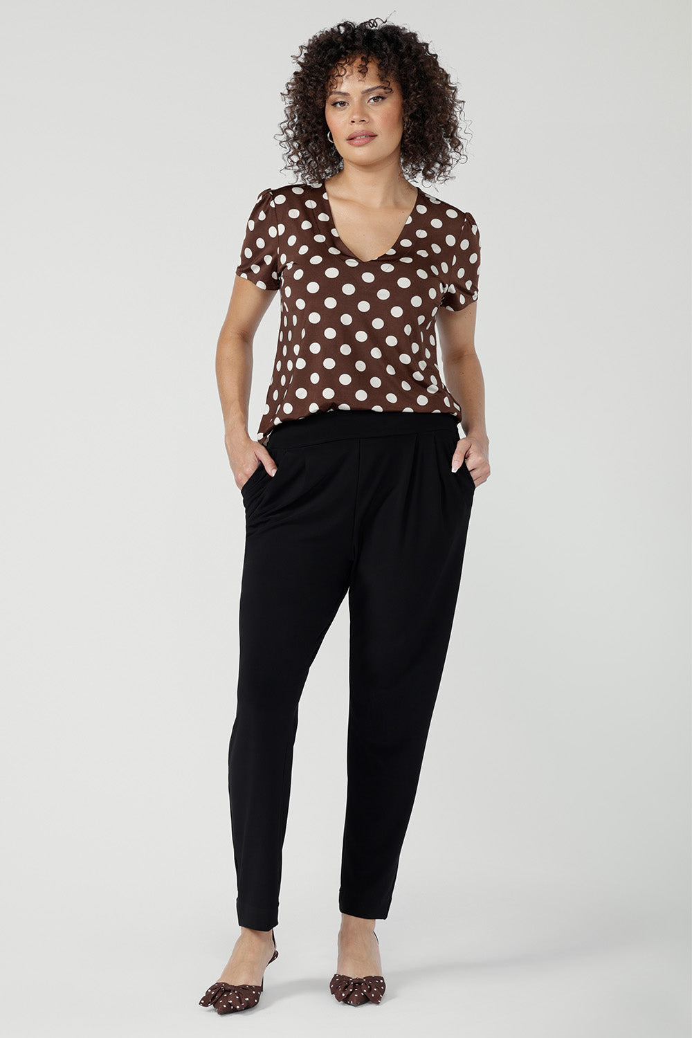 Size 10 woman wearing a brown polka dot top and black work pants with brown spotty heels.