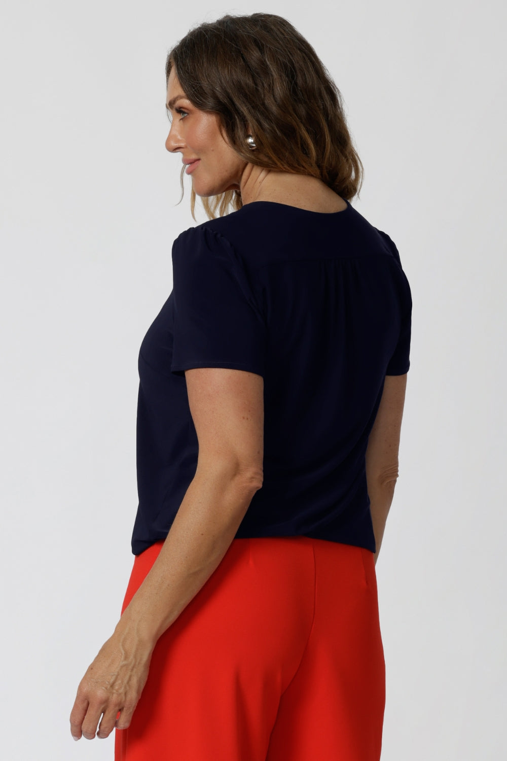 Back view of a good top for over 40s women, a size 12 woman wears a navy jersey top great for corporate wear.  Styled back with a red tailored wide leg pants pants. This tailored top with short sleeves and a v-neckline is made in Australia by Australian fashion brand, Leina & Fleur in sizes 8 to 24.