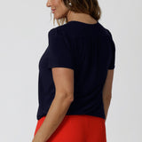 Back view of a good top for over 40s women, a size 12 woman wears a navy jersey top great for corporate wear.  Styled back with a red tailored wide leg pants pants. This tailored top with short sleeves and a v-neckline is made in Australia by Australian fashion brand, Leina & Fleur in sizes 8 to 24.