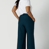 Back view of a size 16 woman wears the Yael pant in Petrol. A high waist tailored pant with tailoring elements like side pockets, zip front and belt loops. Full length wide leg pant suitable for comfortable workwear with a statement Petrol colour. Styled back with white Evan top in white bamboo. Made in Australia for women size 8 - 24.