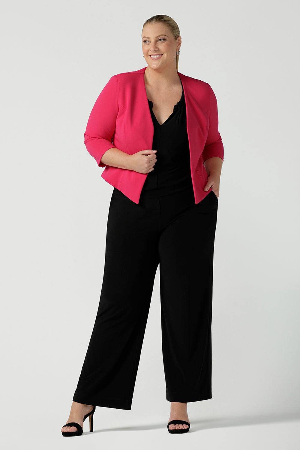 Plus size 18 women wears jumpsuit for curvy women. This black jumpsuit features a halter neck with side pockets and straight legs. Made in Australia in navy stretch jersey this jumpsuit is comfortable for work or weekend wear.  Styled with a pink workwear jacket. 