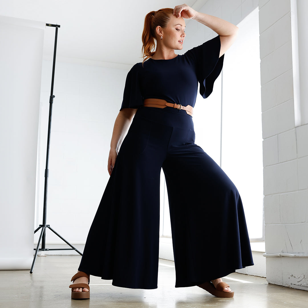 A curvy size 12 woman wears a high neck, navy blue flutter sleeve top tucked in to navy blue, wide leg palazzo pants, all made in Australia by women's clothing brand, Leina & Fleur.