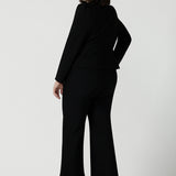 Back view of a Size 10 woman wears the Garcia Jacket in Black. Tailored design with collar and comfortable stretch. Comfortable and stylish workwear for women. Size inclusive fashion and made in Australia for women size 8 - 24. Styled back with Black Kick flare Troy pants, boot leg cut. 