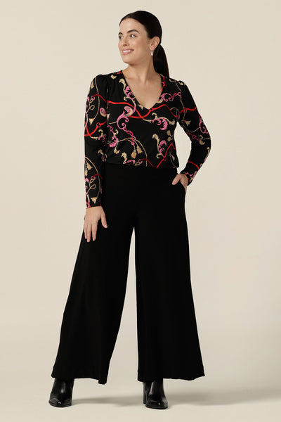 A size 10 woman wears black wide leg pants with pockets, together with a V-neck, long sleeve baroque-print top.  These pull-on, easy care pants are comfortable for your everyday workwear capsule wardrobe. Shop these Australian-made black trousers online in sizes 8 to 24, petite to plus sizes.