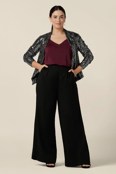 A size 10, petite height woman wears a silver sequin jacket with waterfall front for evening and event wear. Styled with a plum cami top and high-waisted black trousers, this sparkly jacket is glamorous cocktail attire at it's finest! Made in Australia by women's occasionwear brand, Leina & Fleur, shop cocktail jackets online in petite to plus sizes.