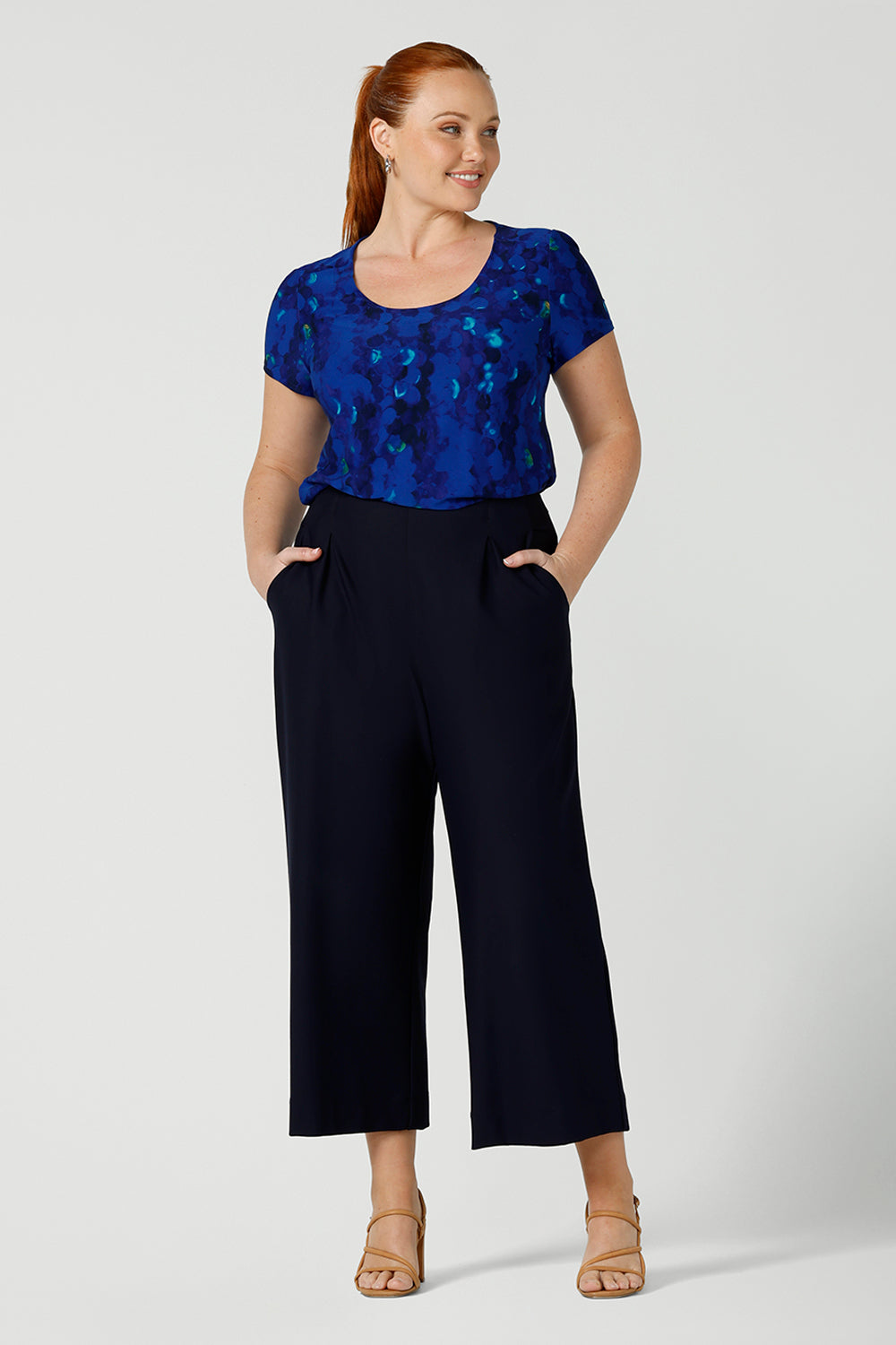 A curvy, size 12 woman wearing a cobalt abstract jersey print, Round neck top with short sleeves with navy wide leg pants. A good top for summer casual wear, or style tucked as a workwear top. Shop made in Australia tops in petite to plus sizes online at Australian fashion brand, Leina & Fleur.