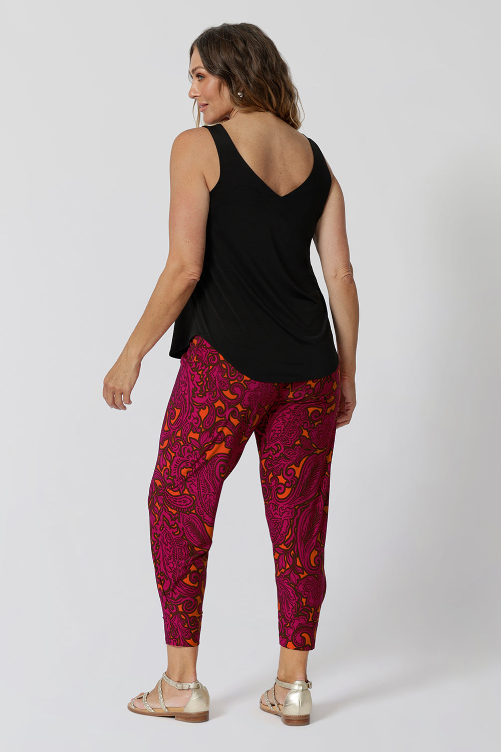 Back view of over 40 year old woman wearing Indi pants in paisley print featuring colours of fuchsia and tangerine. Drop crotch feature provide extreme comfort for busy days and travel adventures. Bold colour way can be styled casually or dressed up for the season.