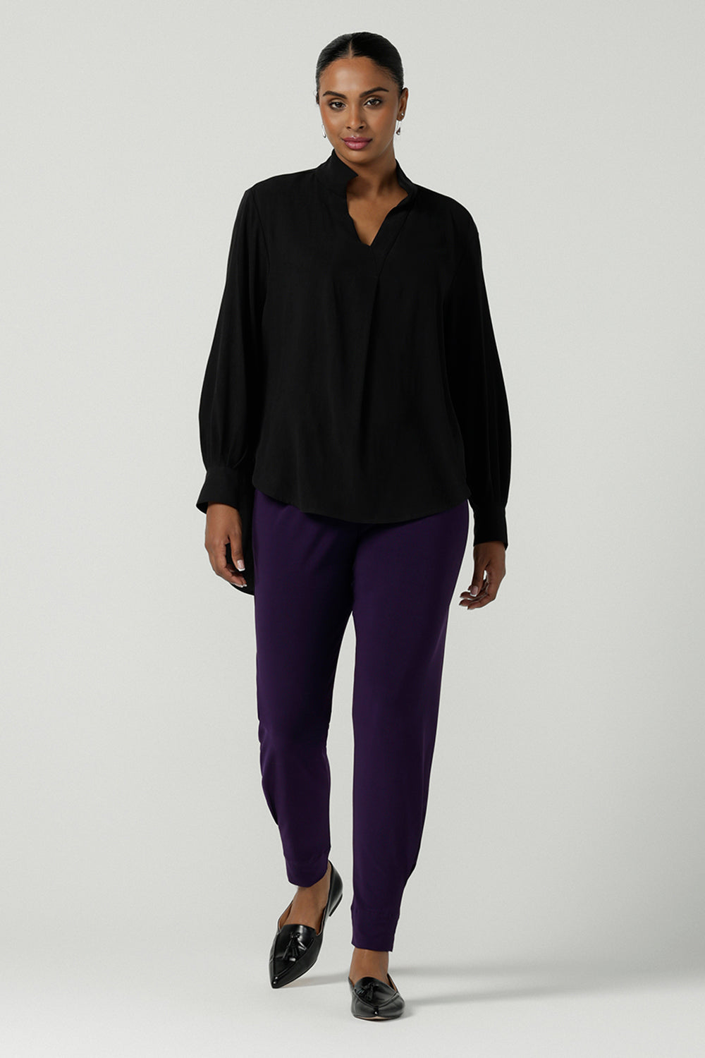 A size 10 woman wearing the Indi Pant in Amethyst. An Indi pant in amethyst with a mid rise waistband, tapered ankle balloon leg style. A made in Australia pant for women. Perfect travel friendly non-crease pant. Size 8-10. Styled back with a black Ellery Shirt in Tencel. 