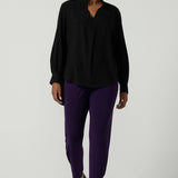 A size 10 woman wearing the Indi Pant in Amethyst. An Indi pant in amethyst with a mid rise waistband, tapered ankle balloon leg style. A made in Australia pant for women. Perfect travel friendly non-crease pant. Size 8-10. Styled back with a black Ellery Shirt in Tencel. 