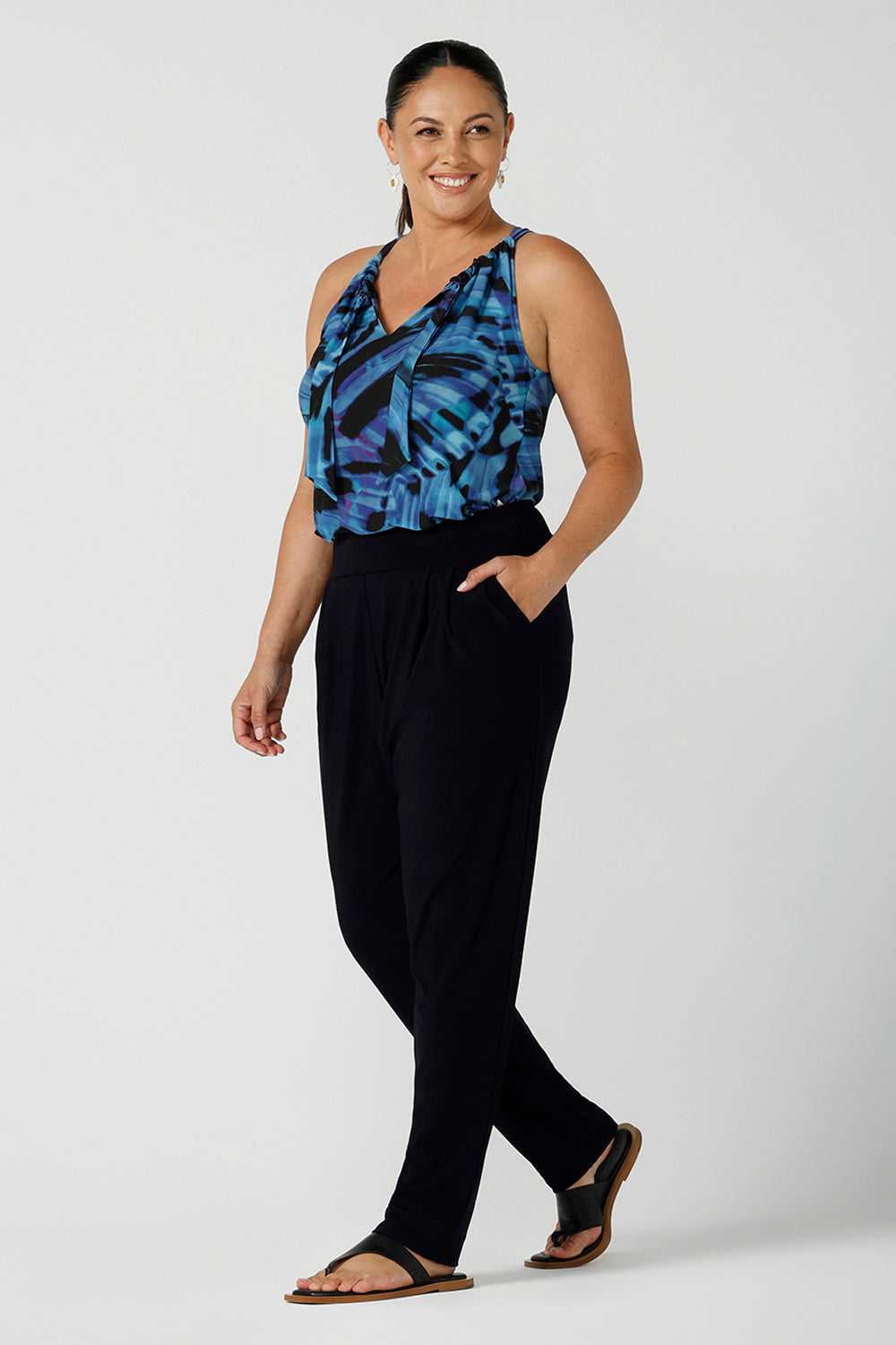 Woman wears tie neck tie top Iris in Flutter with a blue digital print. Styled back with Navy Indi Pants in Jersey.  Size inclusive clothes for women size 8 - 24. Digital blue and purple print. Made in Australia.