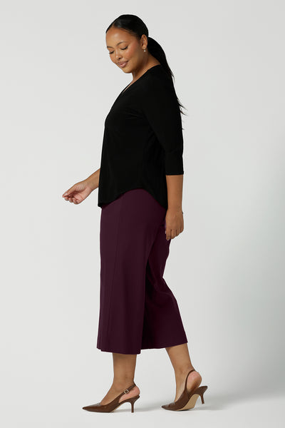 A size 16 curvy woman wears the Jaime top in black. A comfortable corporate work top with soft tailoring elements like a pleat front neckline, V-neck and 3/4 sleeve. Made in Australia for women size 8 - 24. Styled back with plum coloured Bradley culottes. 