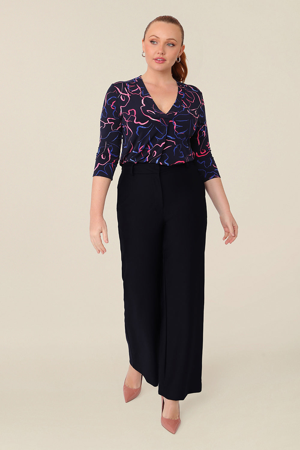 A size 12 woman wears a jersey, V-neck top with 3/4 sleeves. A tailored top for work and corporate wear, the abstract print top is tucked in to wide-leg trousers as a workwear outfit.