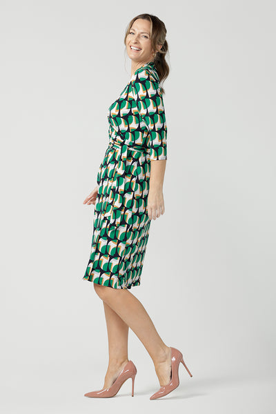 A size 10 woman wears a jersey wrap dress with 3/4 sleeves. In emerald geometric print jersey, this wrap dress is a good corporate dress for work wear capsule wardrobes. Shop Australian-made wrap dresses for work online is sizes 8 to 24.