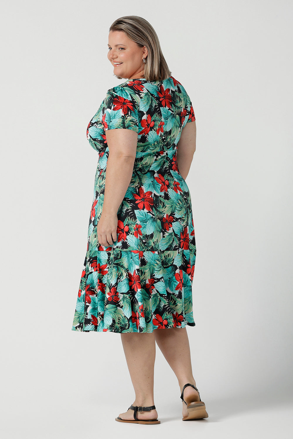 Back view of a size 18 Curvy woman wears the Jillian dress in Havana. A beautiful tropical print with a green leaf print and red flower on a white base that is tropical inspired. A sweetheart neckline style with an empire line. Tier on hem. Made in Australia for women. Size 8 - 24.