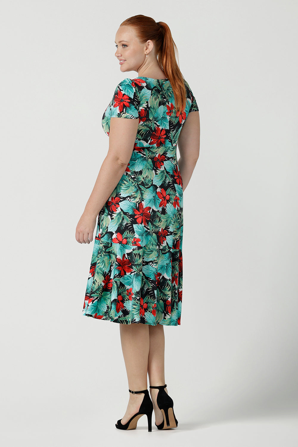 Back view of a size 12 woman wears the Jillian dress in Havana. A beautiful tropical print with a green leaf print and red flower on a white base that is tropical inspired. A sweetheart neckline style with an empire line. Tier on hem. Made in Australia for women. Size 8 - 24.