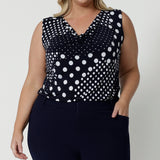 Showing a summer top for curvy women, a size 18, plus size women wears a cowl neck jersey top. This sleeveless top has a navy and white spot print - great for corporate wear as well as casual wear! Made in Australia by Australian and New Zealand clothing brand, Leina & Fleur, shop this top in petite to plus sizes. 