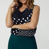 Showing a summer top for over 50s women, a size 12 woman wears a cowl neck jersey top. This sleeveless top has a navy and white spot print - great for corporate wear as well as casual wear! Made in Australia by Australian and New Zealand clothing brand, Leina & Fleur, shop this top in petite to plus sizes. 