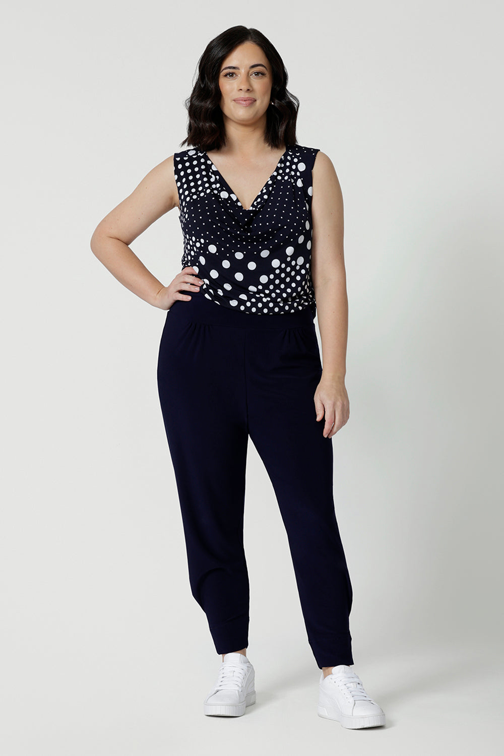 Showing a summer top for petite women, a size 12 woman wears a cowl neck jersey top. This sleeveless top has a navy and white spot print -and is worn with navy travel pants for a travel outfit look! Made in Australia by Australian and New Zealand clothing brand, Leina & Fleur, shop this top in petite to plus sizes. 