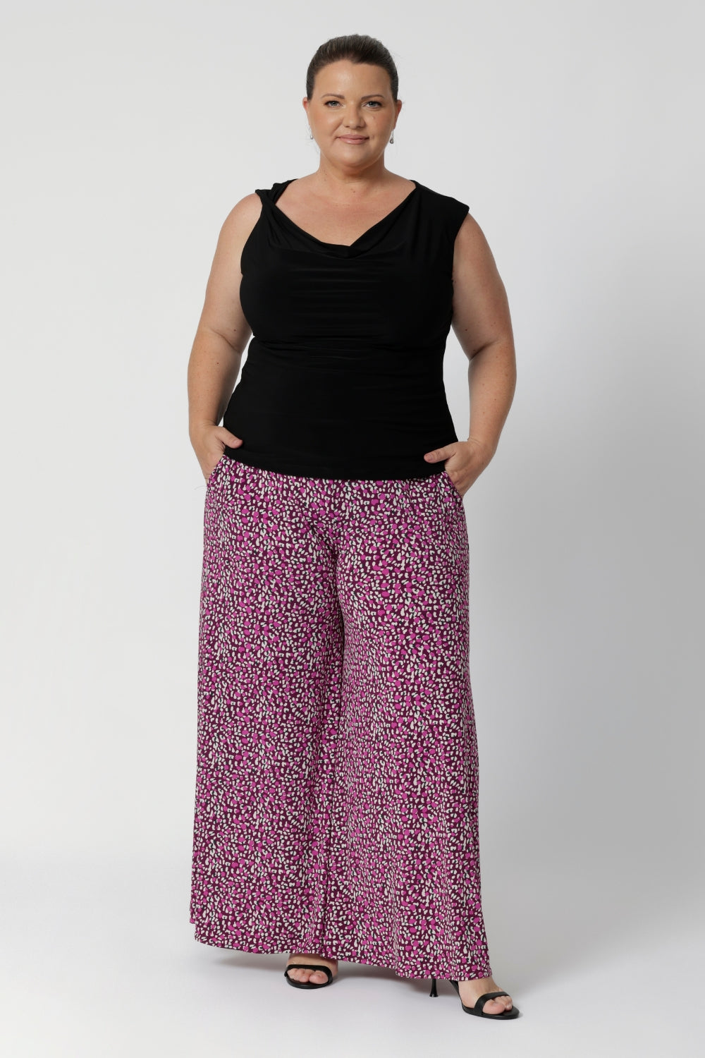 This reversible double-layer top offers two chic styling options: a cowl neckline with a twisted shoulder detail, or a more structured boat-neck look for versatility. Plus size woman wears black reversible top with printed pants and black heels for a possible workwear or eventing look.