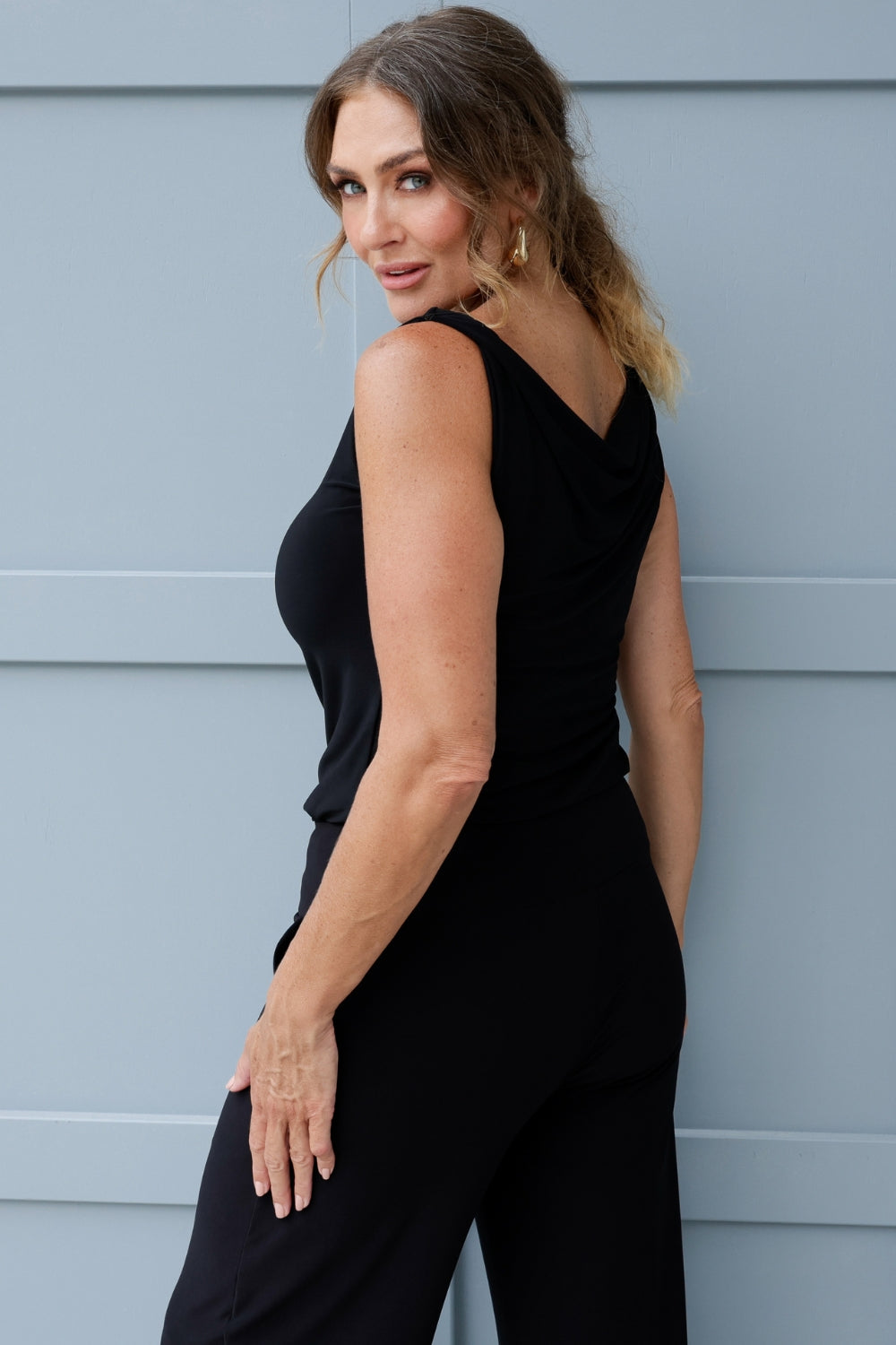 Black reversible top offers two chic styling options, a cowl neckline with a twisted shoulder detail, or a more structured boat-neck look for versatility. Image shows size 12 model wearing her Jordan Top in black with black wide leg pants. 