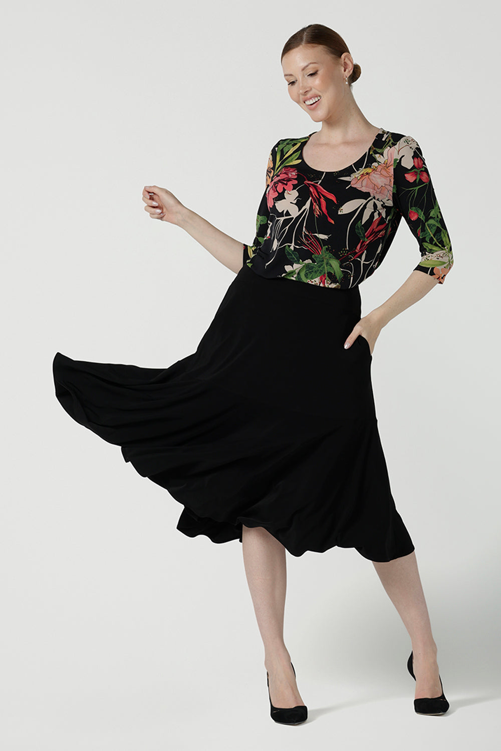 A size 10 woman wears the Jules top in Bloomsbury print. A curved neckline style with 3/4 sleeves. Made in easy care jersey fabric with a beautiful floral print with red, pink, orange, green and vanilla on a black base. Made in Australia for women size 8-24. Styled back with the Berit skirt in black.