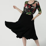 A size 10 woman wears the Jules top in Bloomsbury print. A curved neckline style with 3/4 sleeves. Made in easy care jersey fabric with a beautiful floral print with red, pink, orange, green and vanilla on a black base. Made in Australia for women size 8-24. Styled back with the Berit skirt in black.