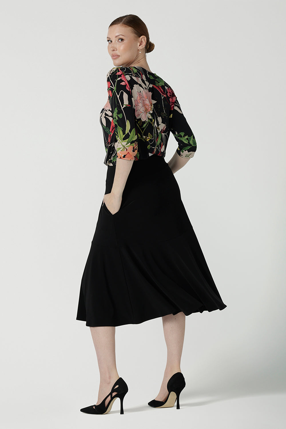 A size 10 woman wears the Jules top in Bloomsbury print. A curved neckline style with 3/4 sleeves. Made in easy care jersey fabric with a beautiful floral print with red, pink, orange, green and vanilla on a black base. Made in Australia for women size 8-24. Styled back with the Berit skirt in black.